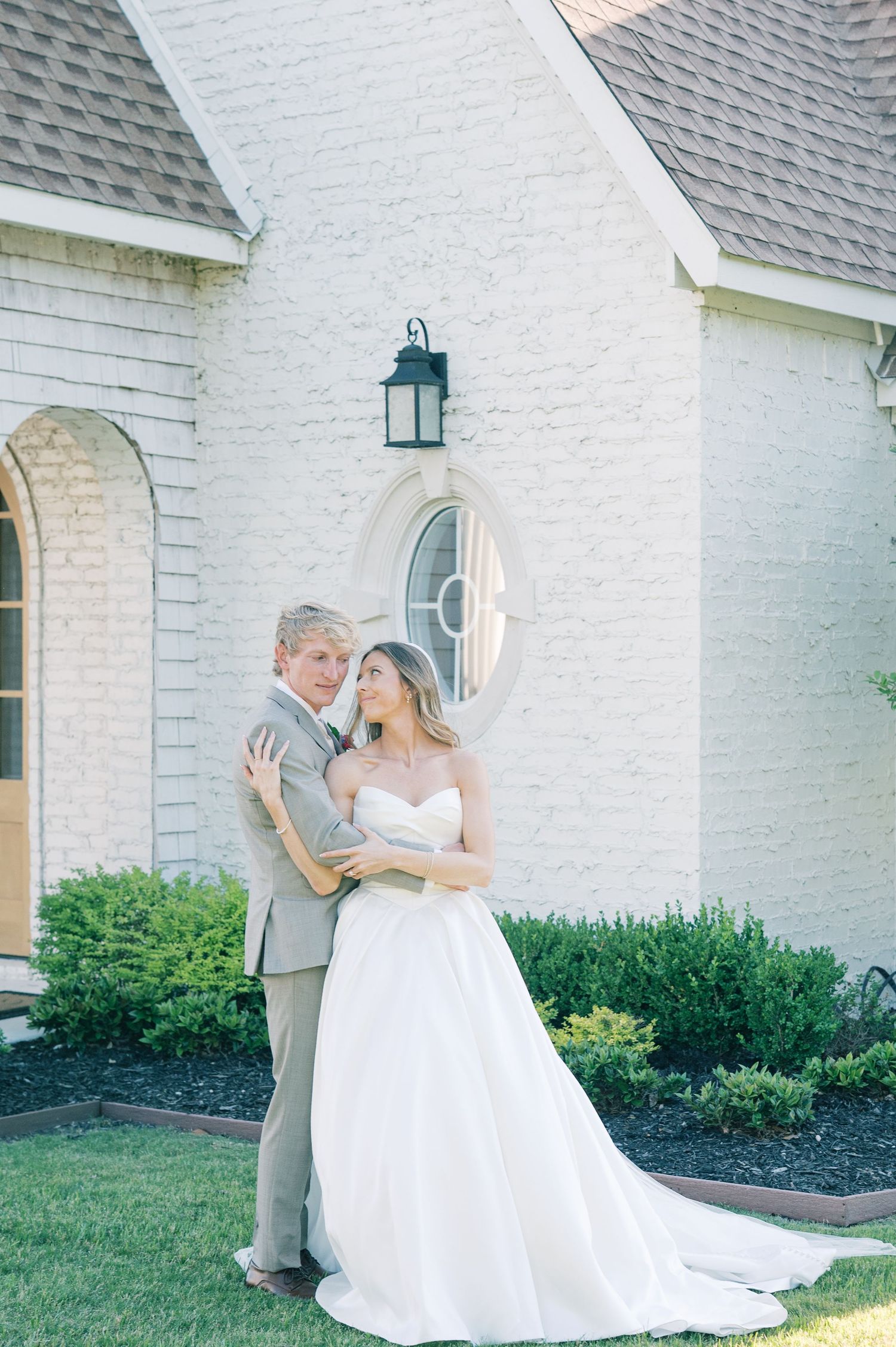 Bride and groom embracing outside a white brick chapel, bride in strapless ball gown with train, groom in gray suit.