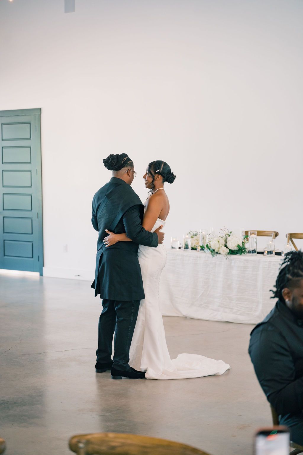 Bride and groom share an intimate first dance in a bright white wedding venue near a draped sweetheart table.