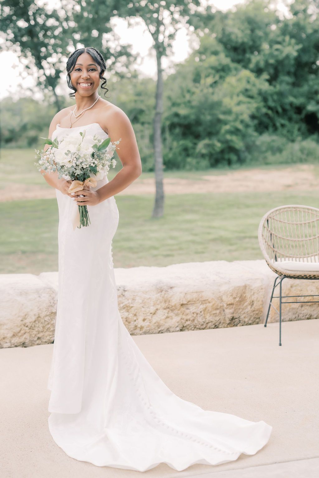 Smiling bride in strapless white gown holding white floral bouquet, standing outdoors on stone patio with green trees.
