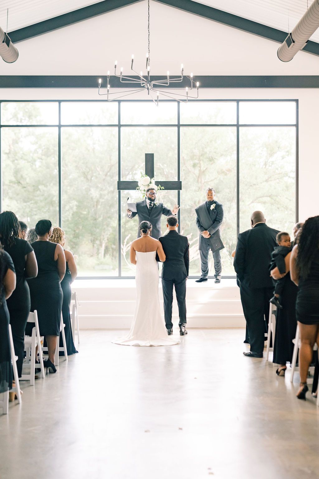 Bride and groom stand at the altar during an elegant indoor wedding ceremony with large windows.