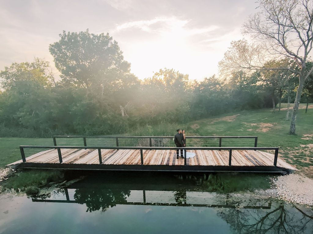 Person standing on a wooden dock over a calm pond at sunset, surrounded by lush green trees in a serene landscape.