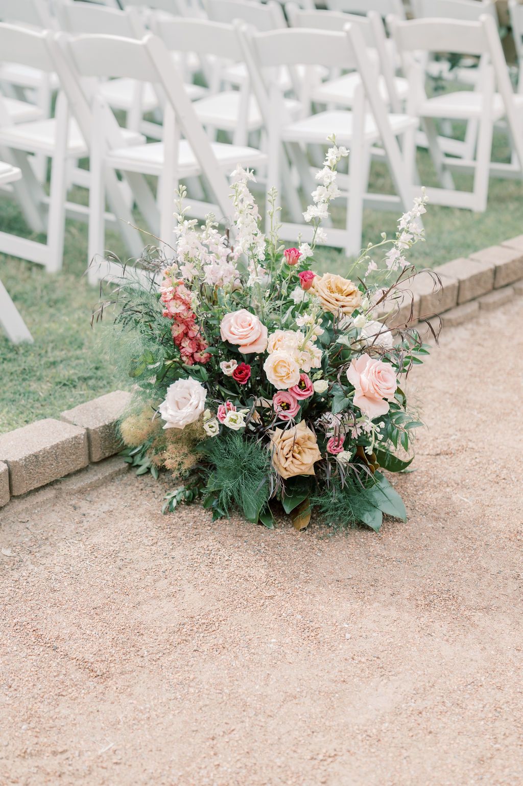 Lush floral arrangement with pink, peach roses and greenery placed along wedding ceremony aisle with white chairs.
