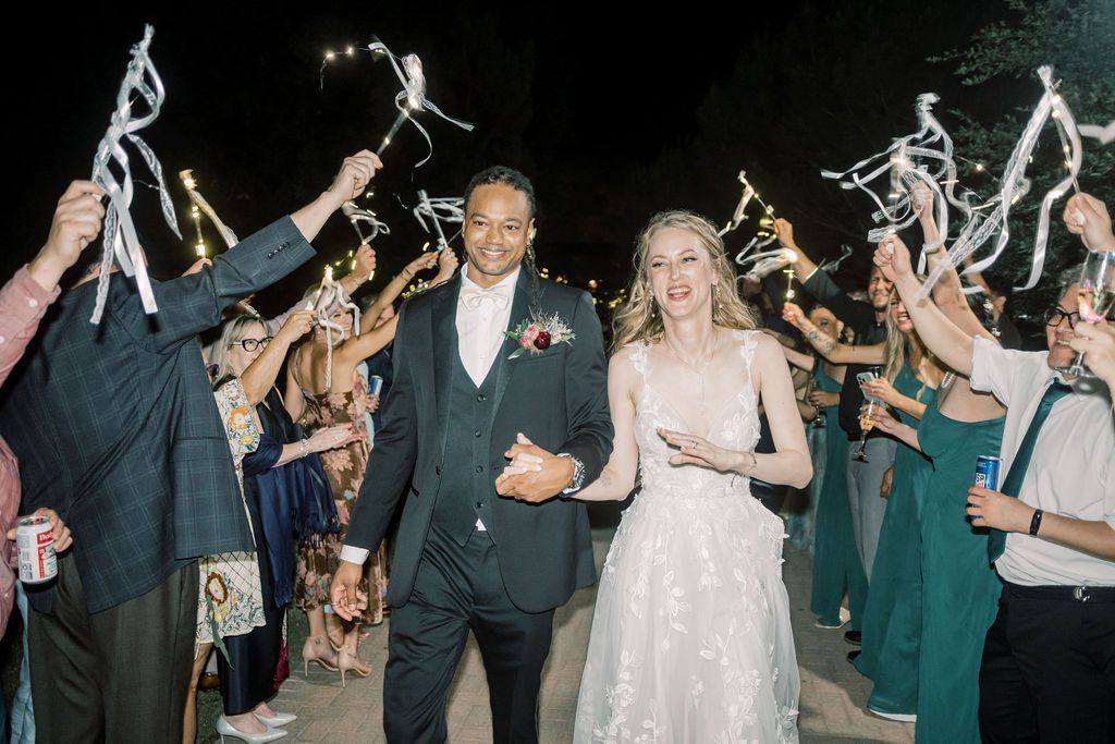 Bride and groom walk through sparkler send-off tunnel with cheering guests at outdoor evening wedding reception.