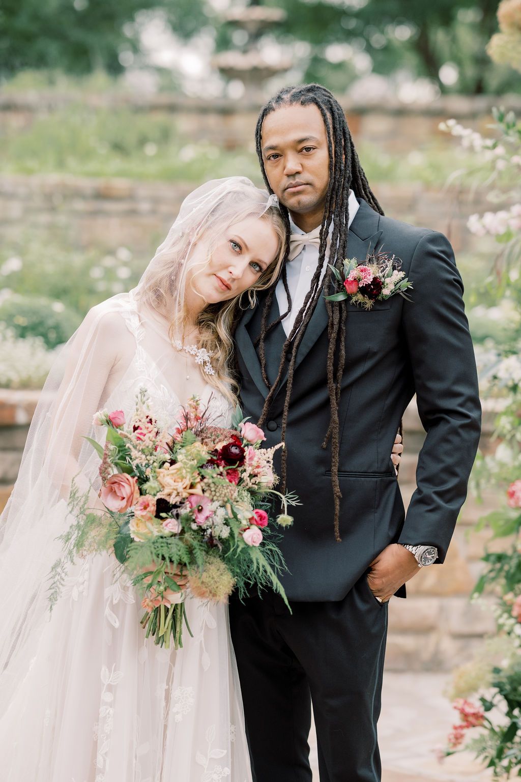 Interracial couple in wedding attire pose in garden, bride holds colorful bouquet, groom wears black suit.