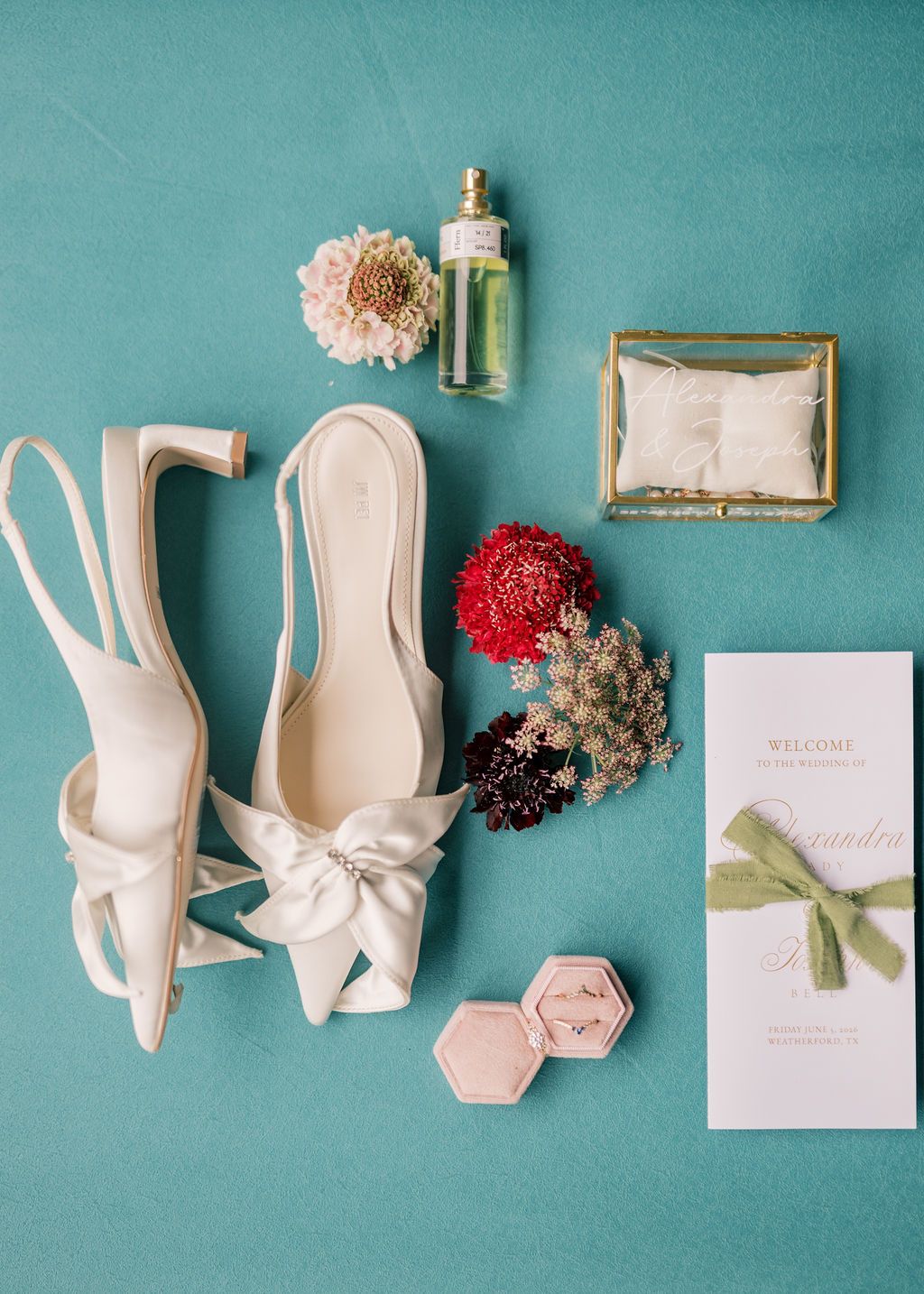Elegant bridal flat lay with ivory bow heels, perfume, ring box, dried florals, and wedding stationery on teal background.