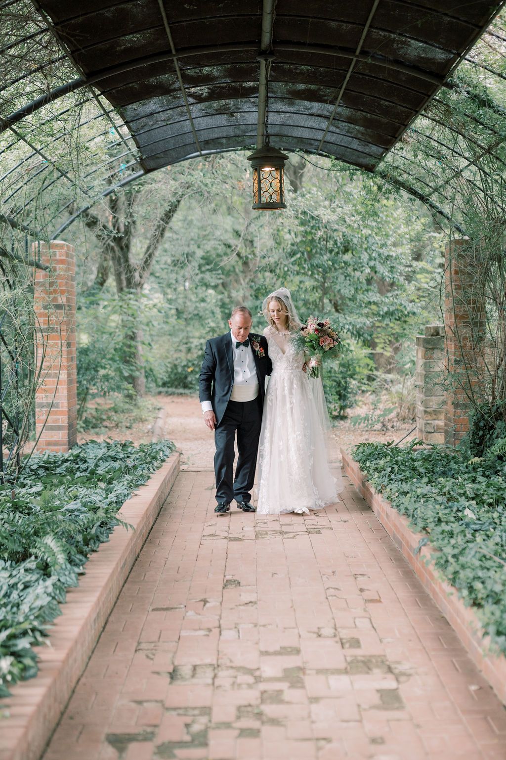 Bride and groom walk hand in hand through a lush garden pergola pathway on their wedding day.