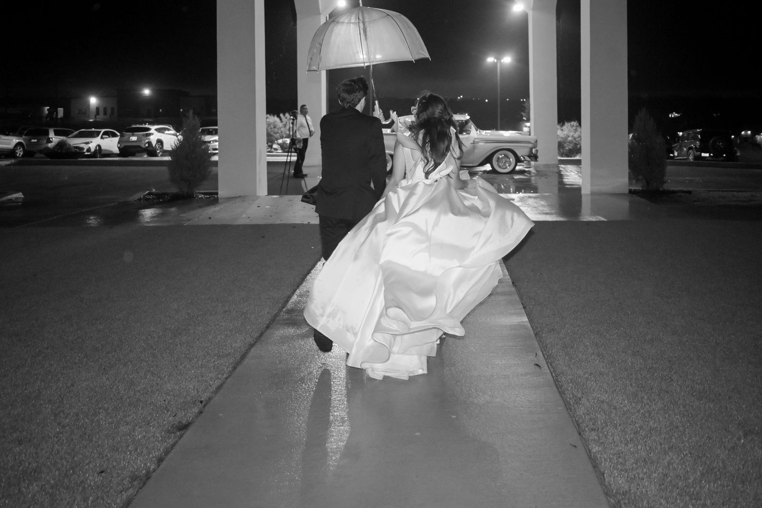 Black and white photo of a bride and groom walking under an umbrella in the rain at night.