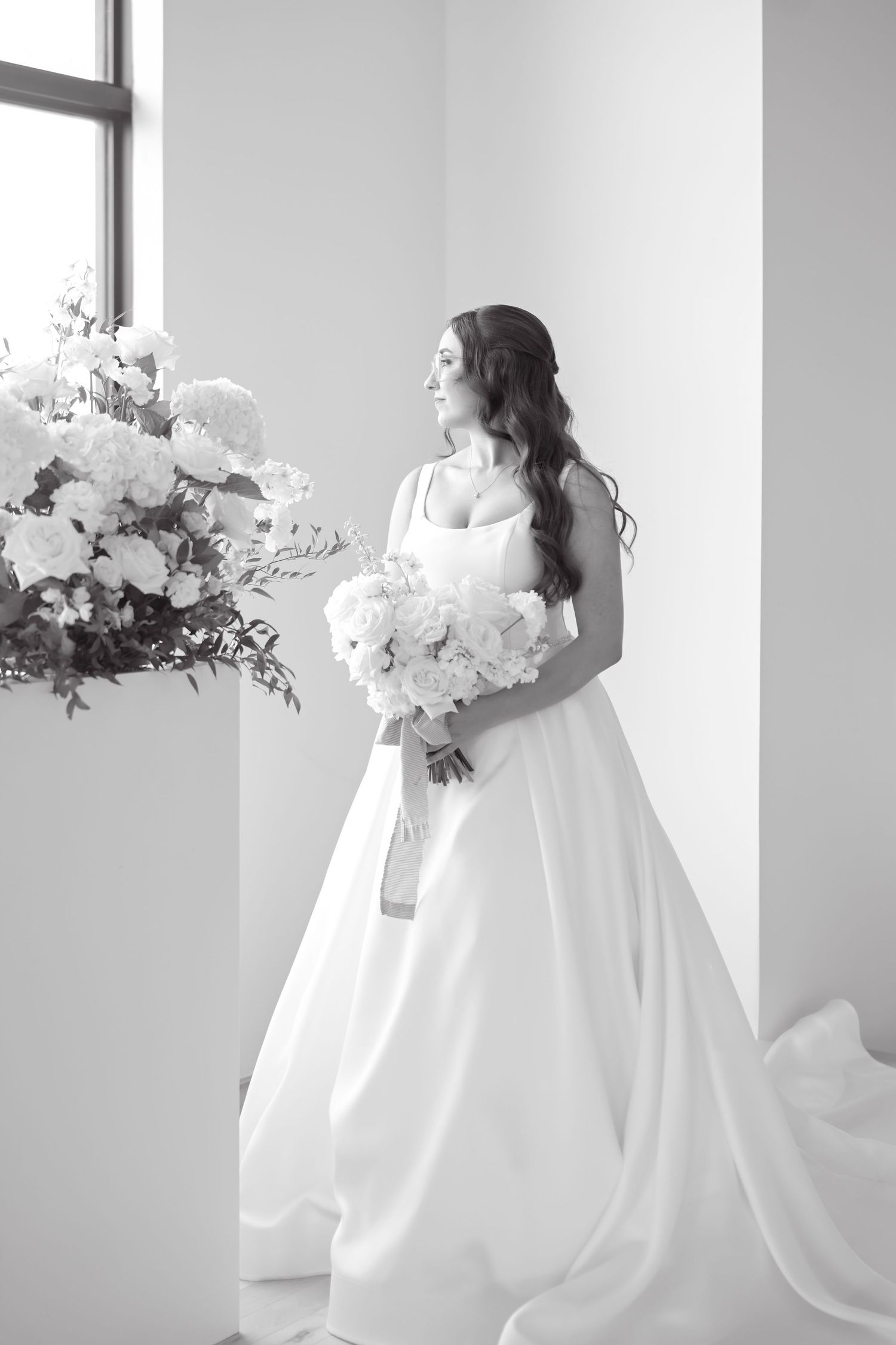 Elegant bride in a white ball gown holding a floral bouquet standing near large blooms in a bright studio.