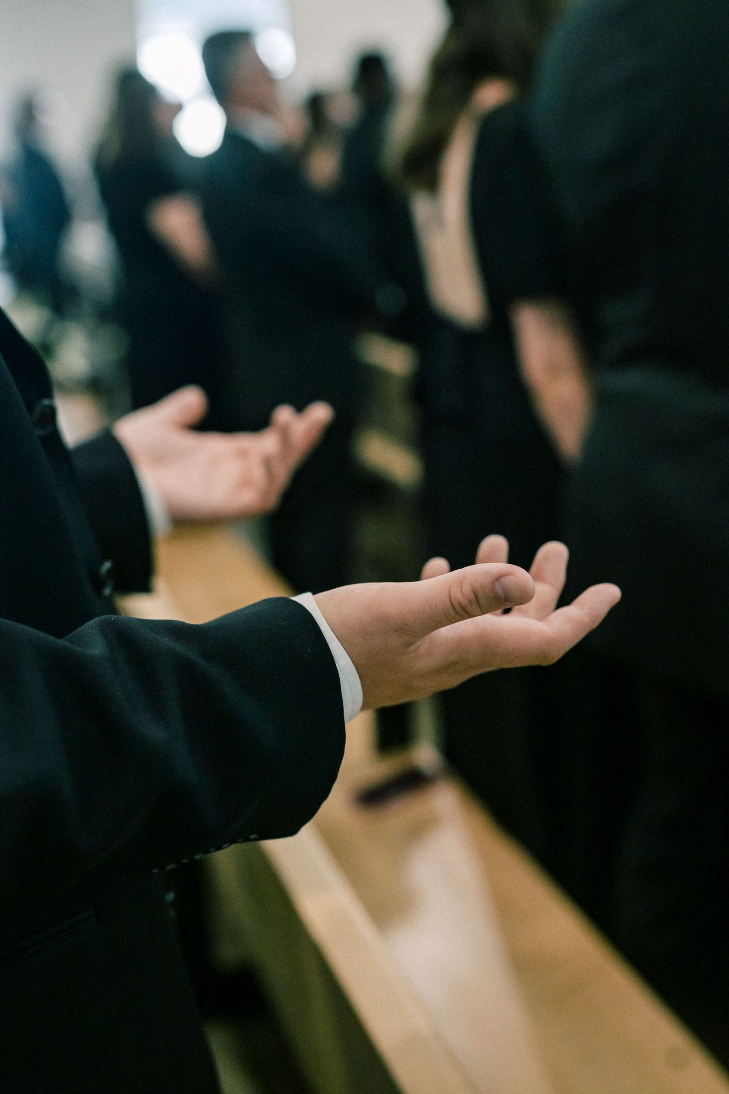 Wedding guests in black formal attire hold open hands in prayer during a religious ceremony beside wooden pews.