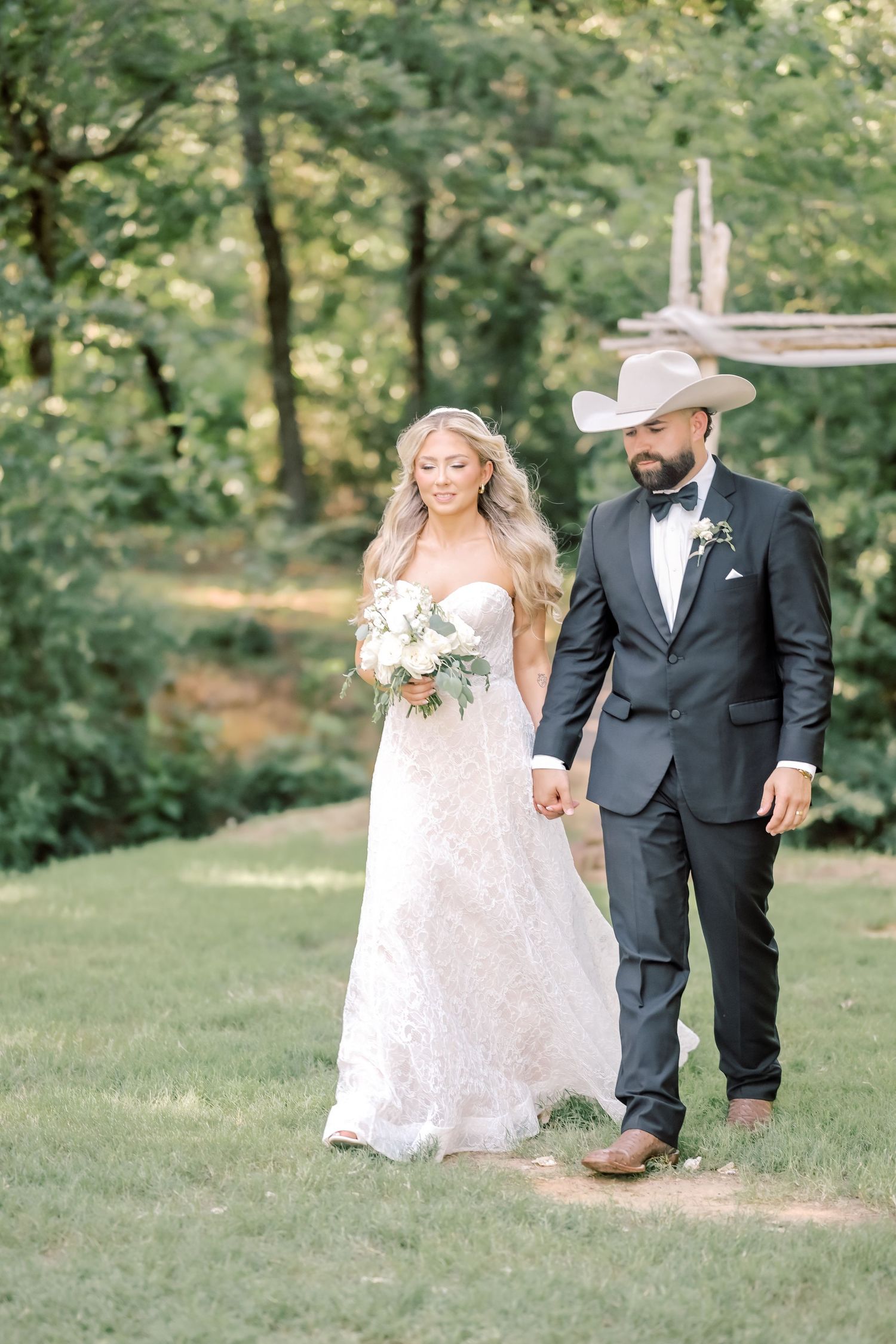 Bride in lace gown and groom in black suit with cowboy hat walk hand in hand outdoors.