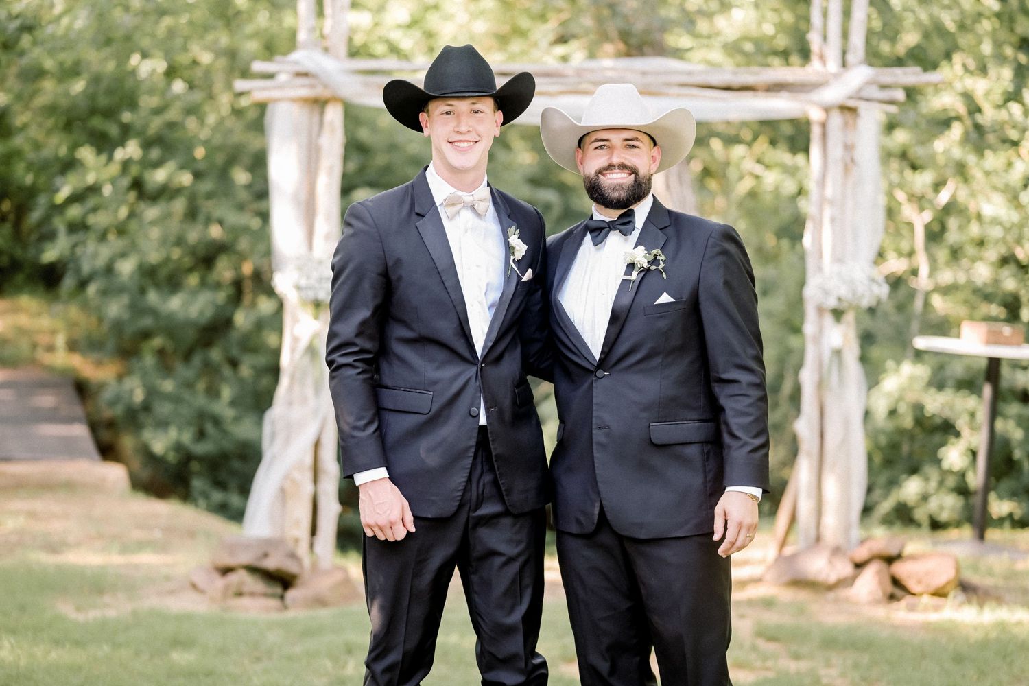 Two groomsmen in black suits and cowboy hats smile at rustic outdoor wedding ceremony.