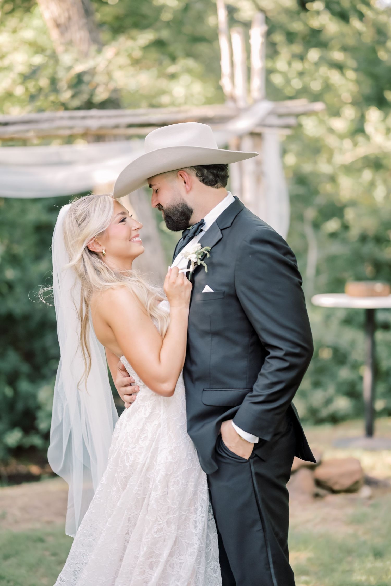 Bride in lace gown smiles at bearded groom wearing tan cowboy hat during romantic outdoor wedding portraits.
