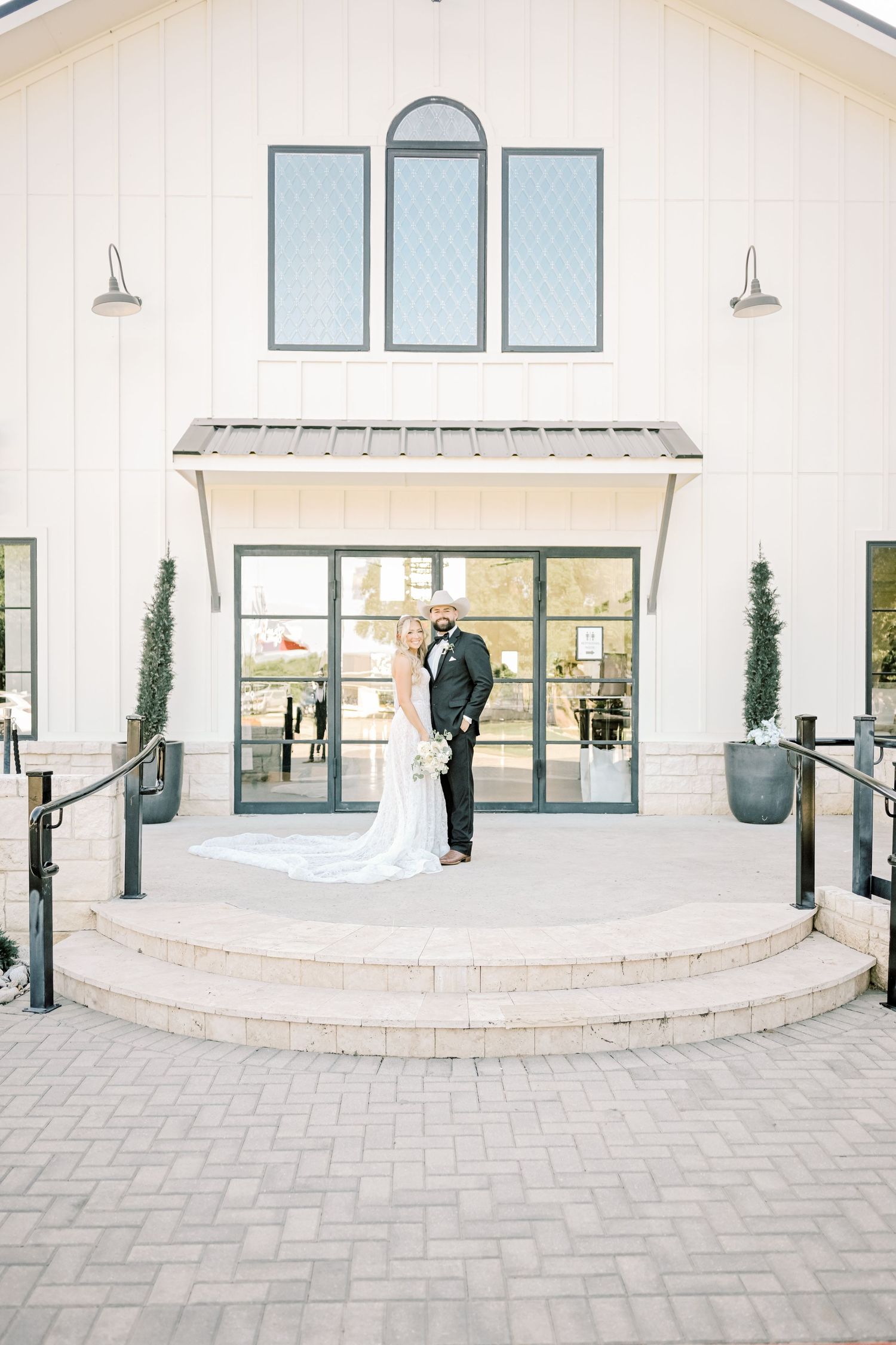 Bride and groom pose on steps outside a white modern farmhouse wedding venue.