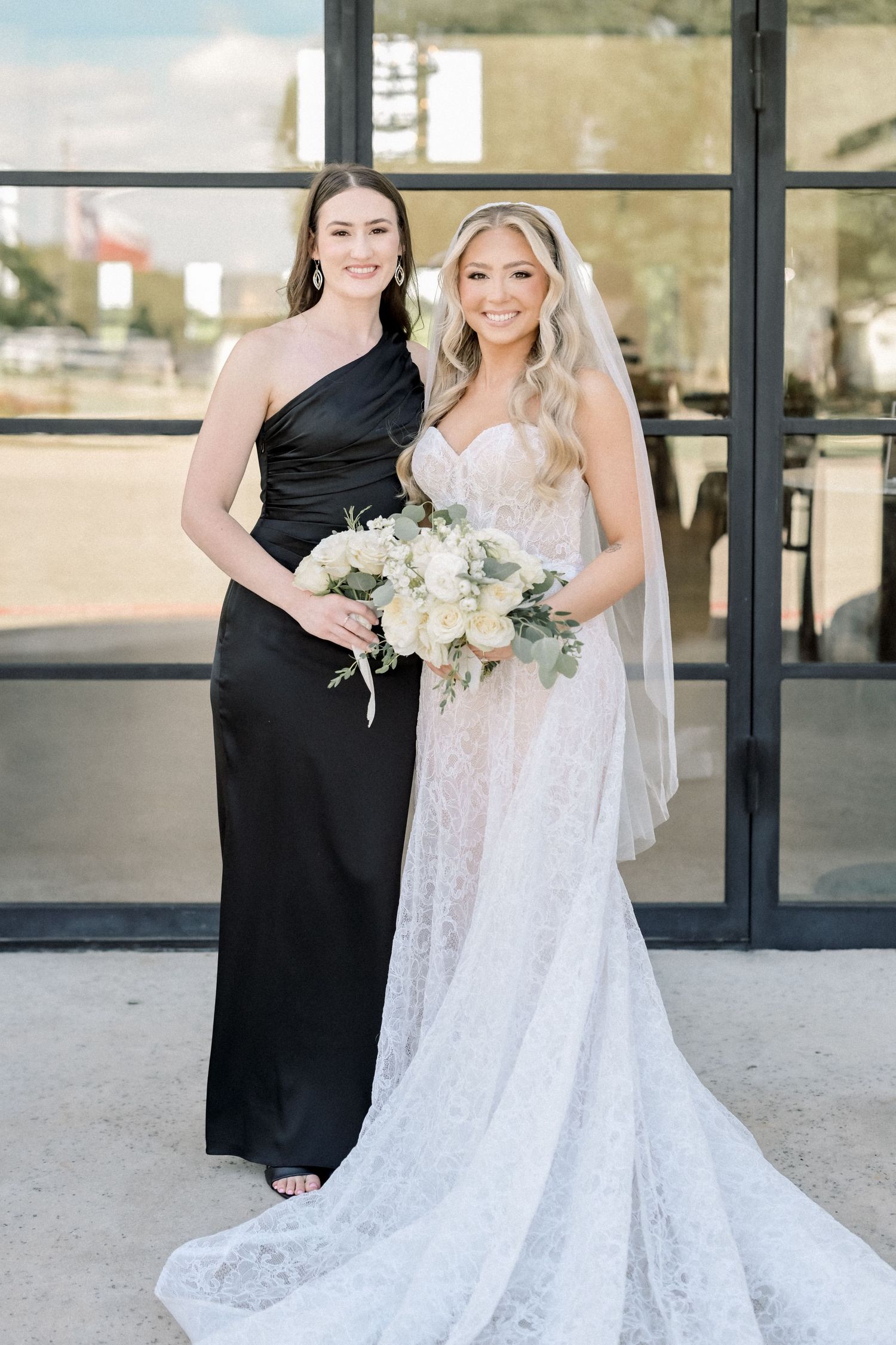 Bridesmaid in black one-shoulder gown standing with blonde bride in lace wedding dress outdoors.