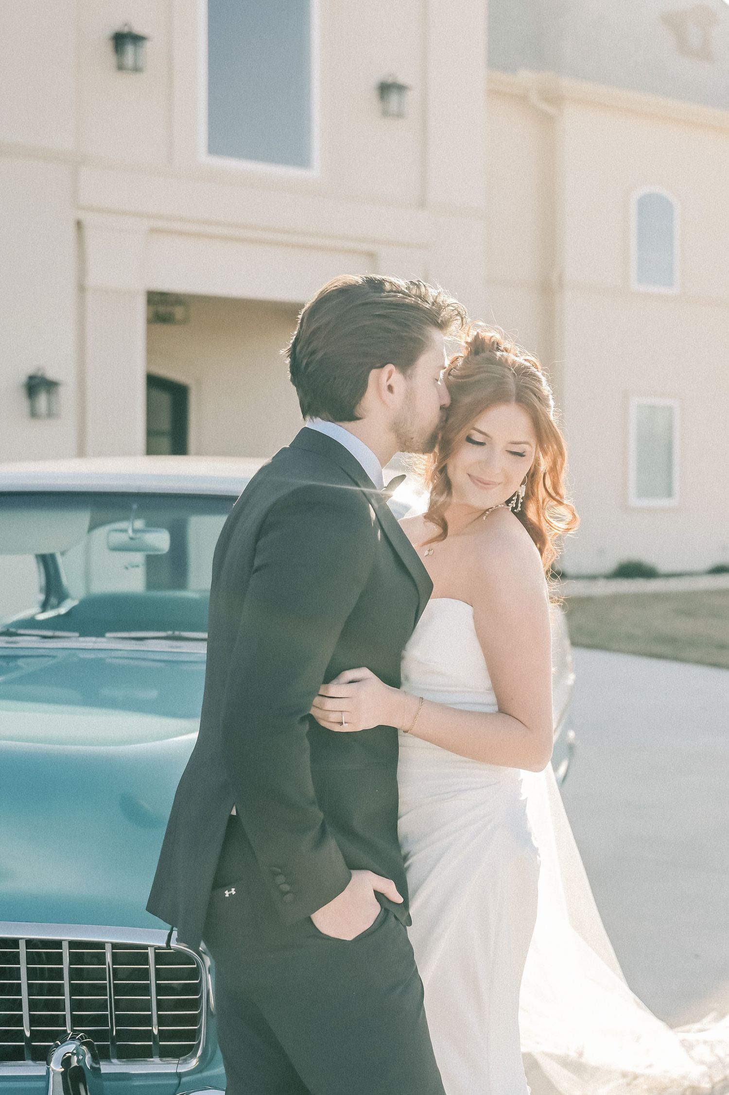 A romantic moment between newlyweds embracing outdoors in soft lighting with a vintage car in the background.
