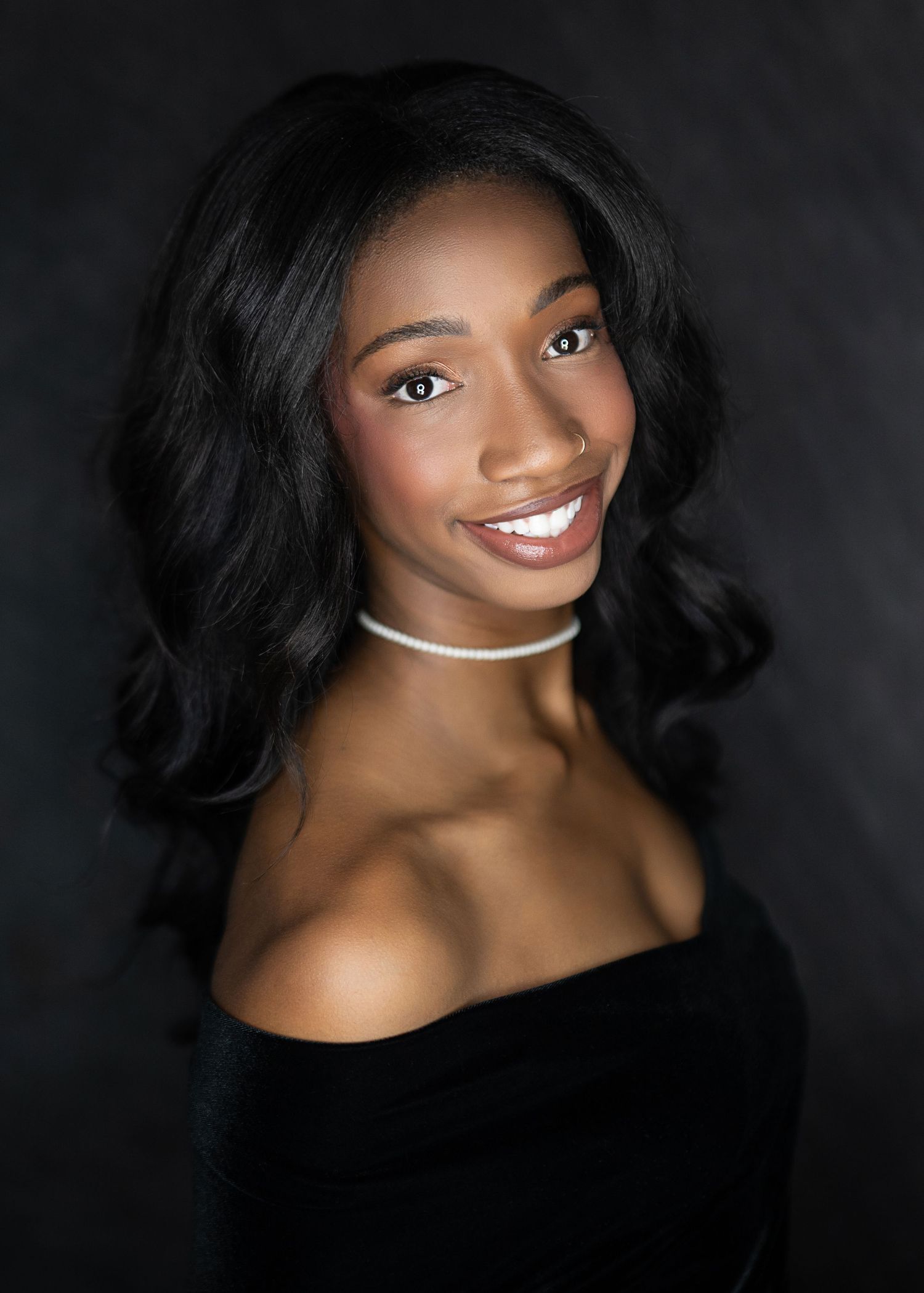 Young woman in black off-shoulder top and white choker smiles confidently against dark studio background.