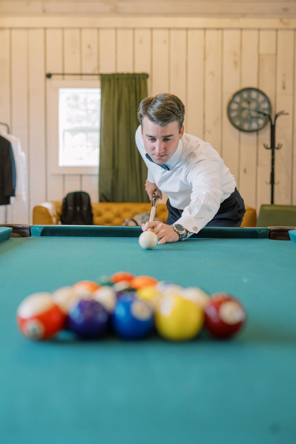A man in a white dress shirt leans over a pool table, aiming his cue at billiard balls on green felt.