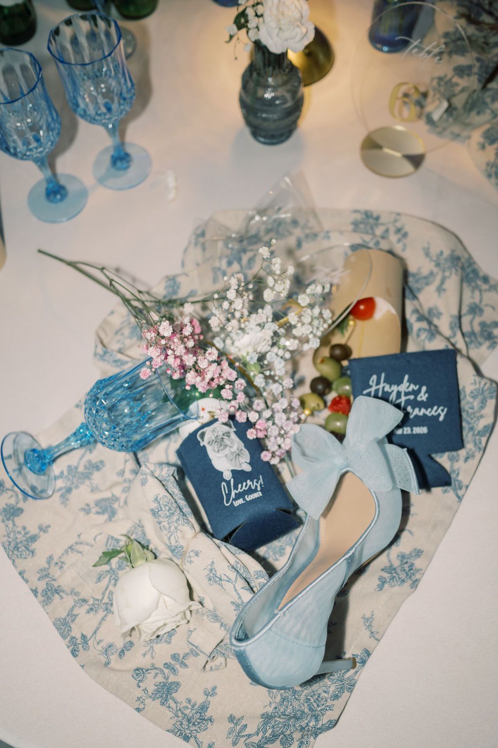 Blue wedding flat lay with toile fabric, floral koozies, baby's breath, blue heels, and decorative glasses.