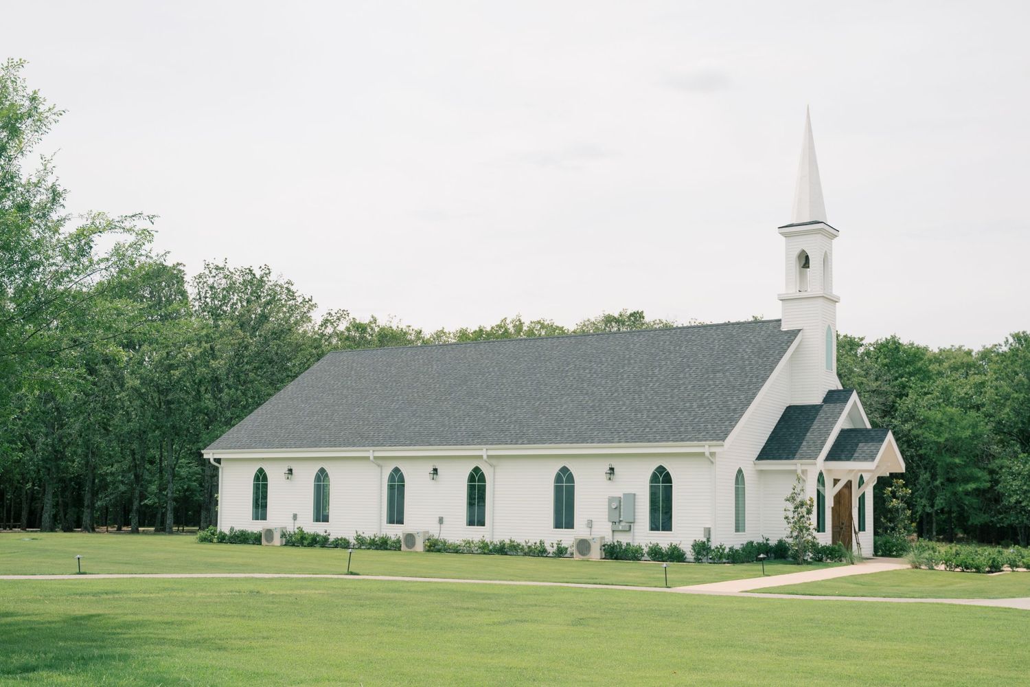 White chapel wedding venue with steeple surrounded by lush green trees and manicured lawn.