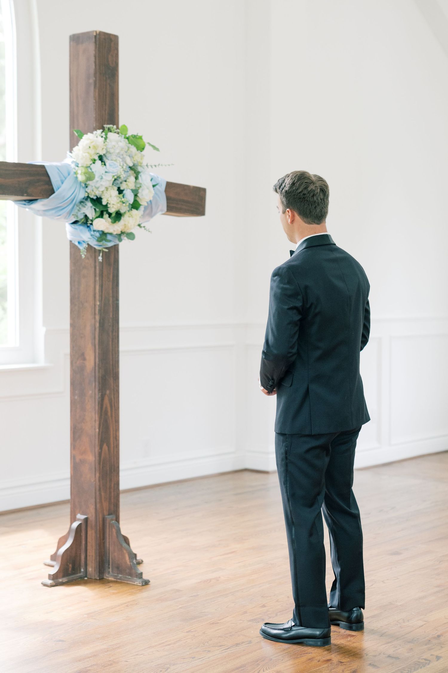 Groom in black suit standing before a wooden cross adorned with a white floral arrangement in a chapel.