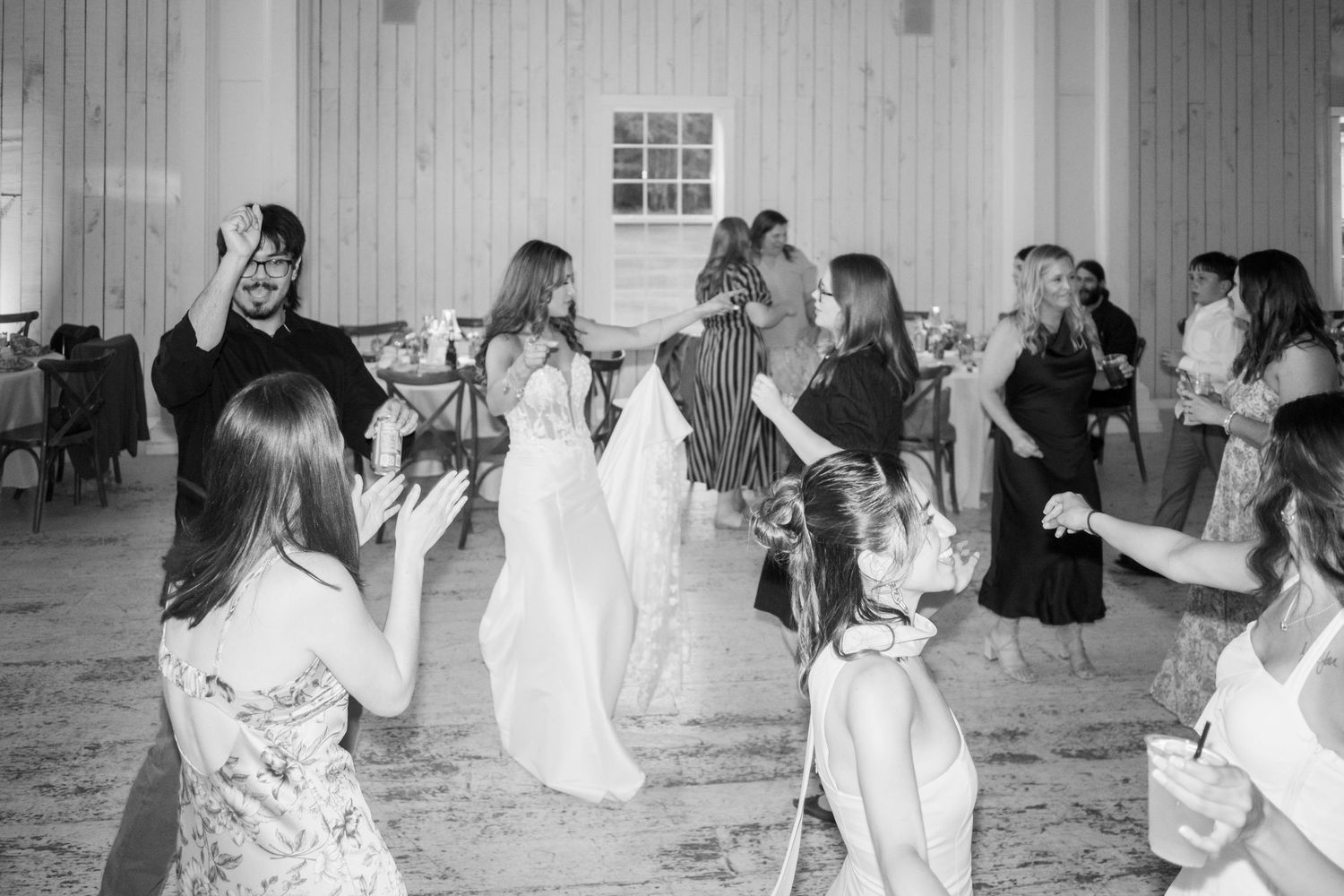Guests dance joyfully at a wedding reception in a white barn venue, captured in black and white.