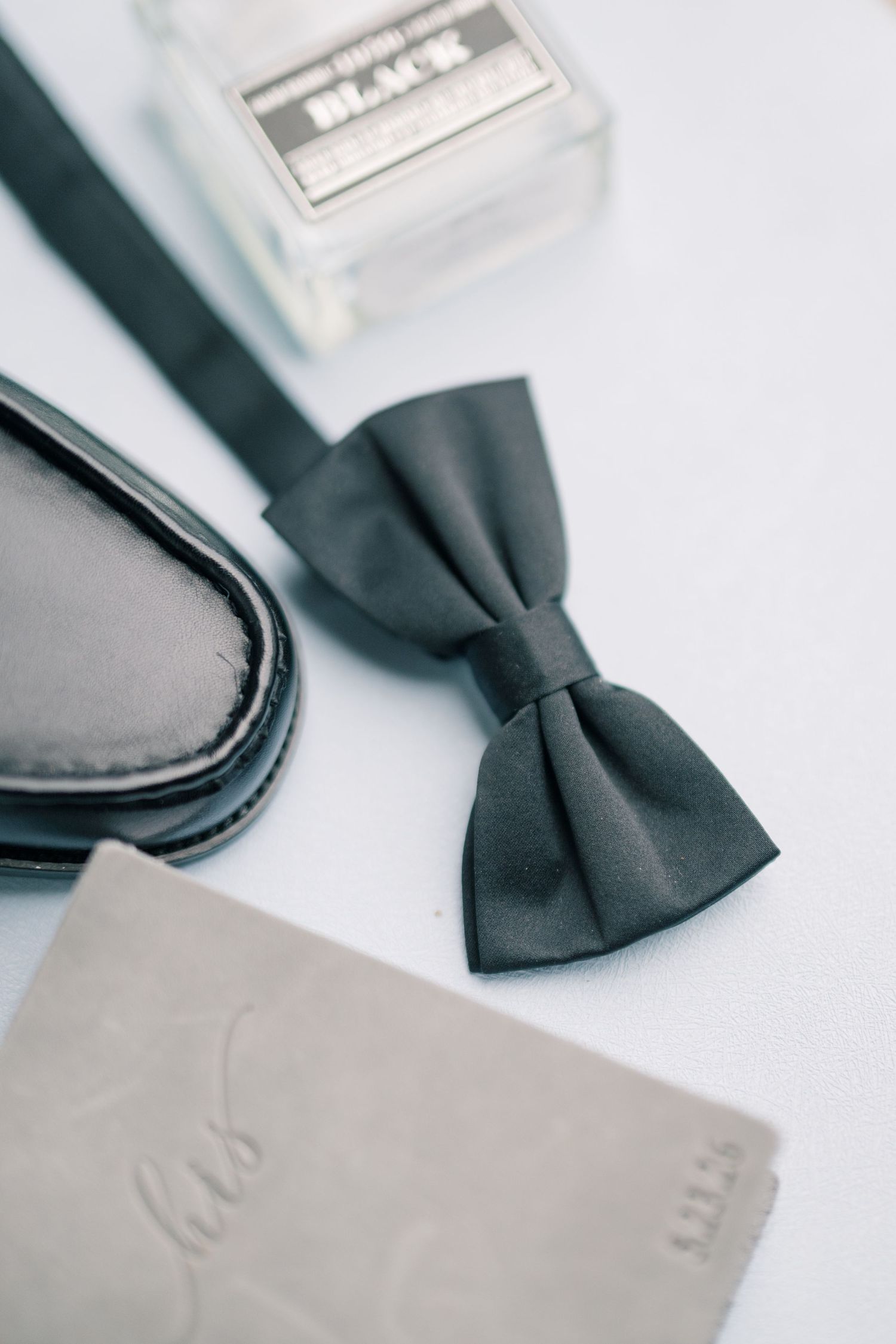 Black bow tie, dress shoes, cologne, and wallet arranged on white surface for wedding day groom details.