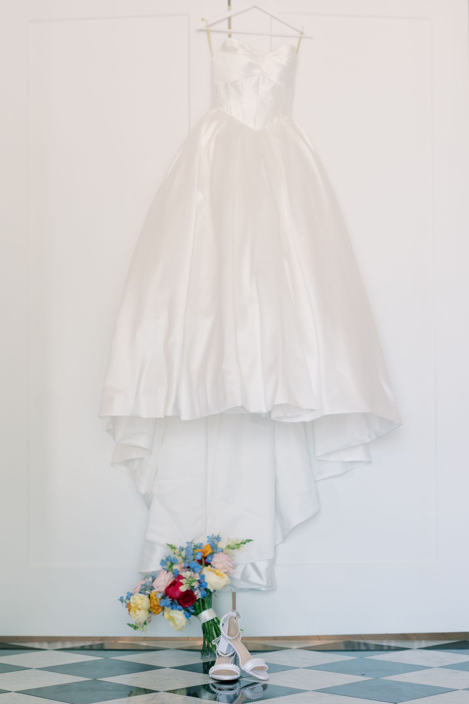 White ball gown wedding dress hanging on wall with colorful bridal bouquet and white heels below.