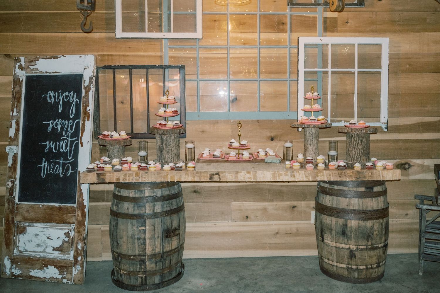 Rustic wedding dessert table displayed on wooden barrels with cupcake tower and chalkboard sign in barn venue.