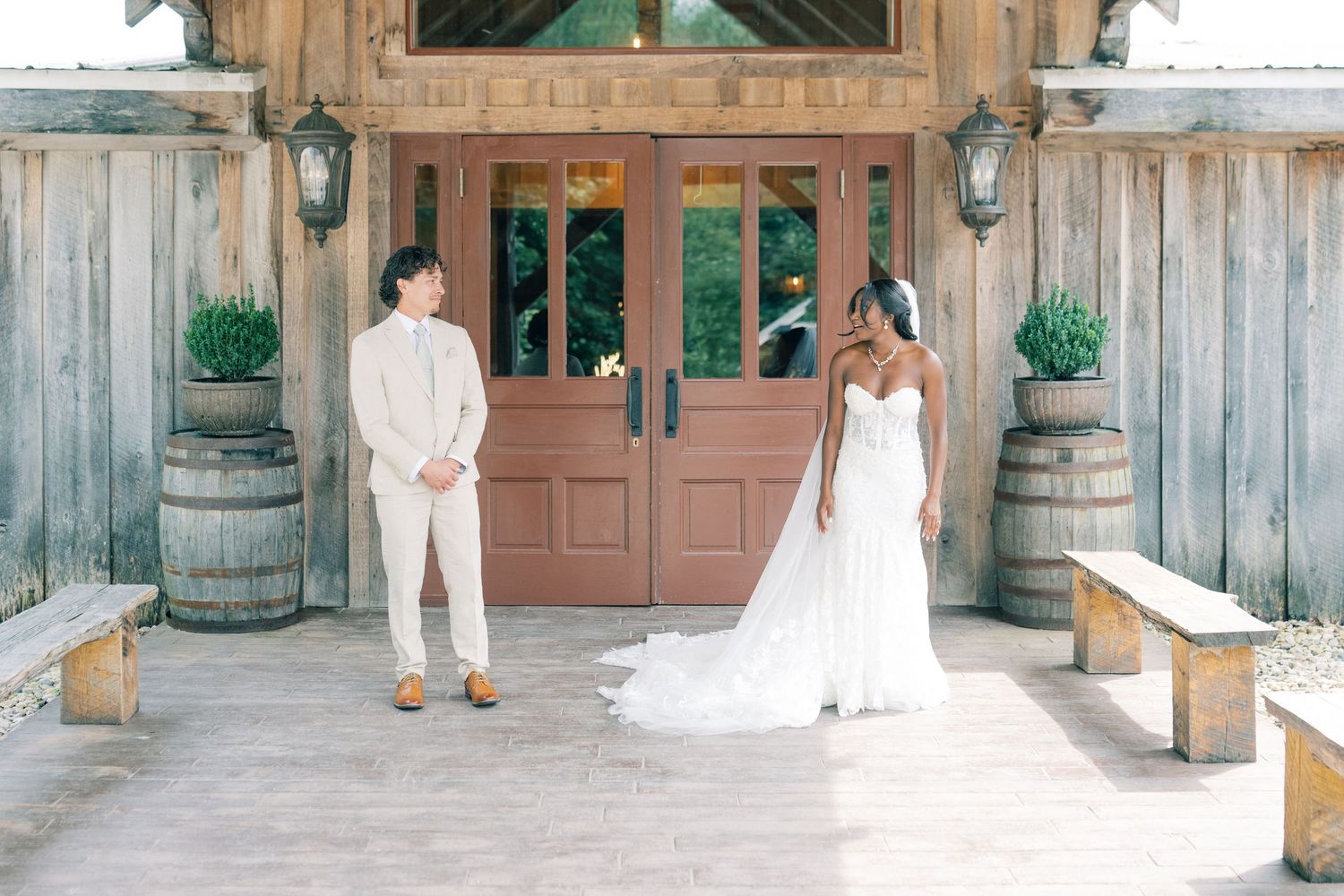 Bride in white gown and groom in beige suit stand before rustic wooden barn doors with wine barrel decor.