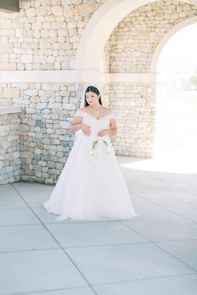 Elegant bride in flowing white gown standing beneath stone archway in natural light.