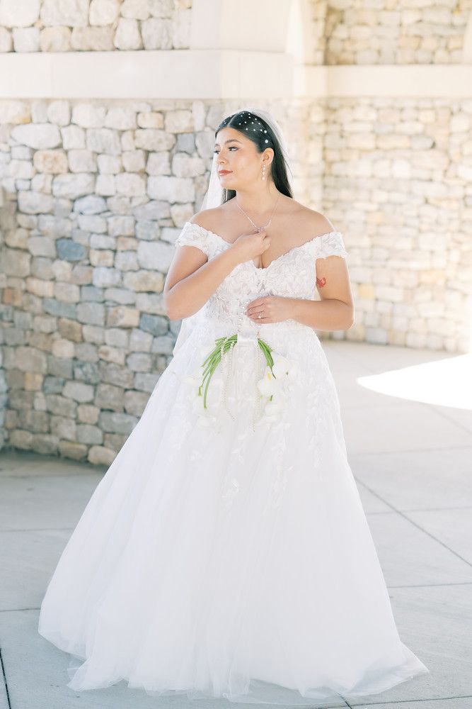 An elegant white wedding dress with floral details photographed against a stone wall backdrop.