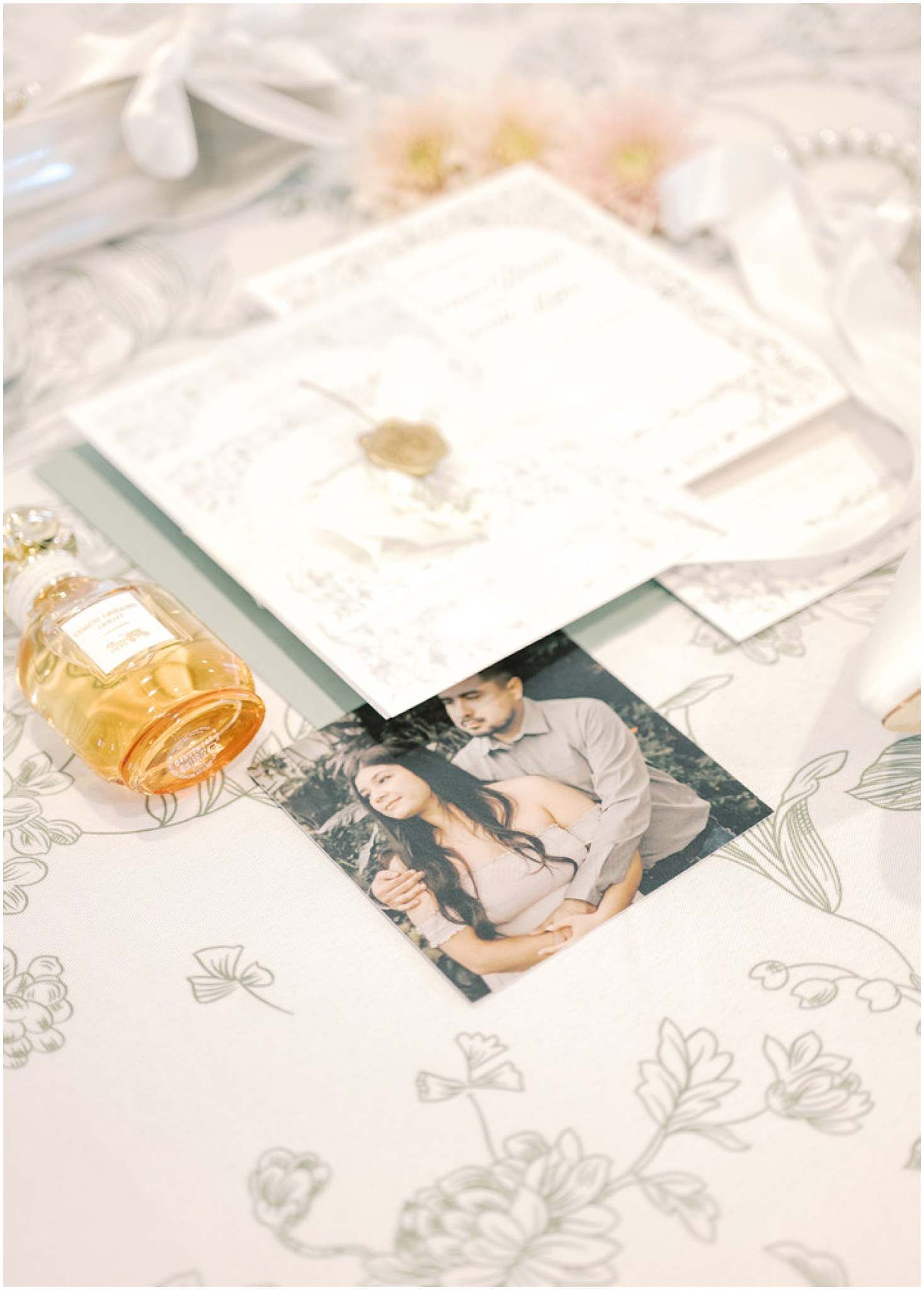 Wedding details flatlay with photographs, floral stationery and perfume bottle on delicate patterned surface.