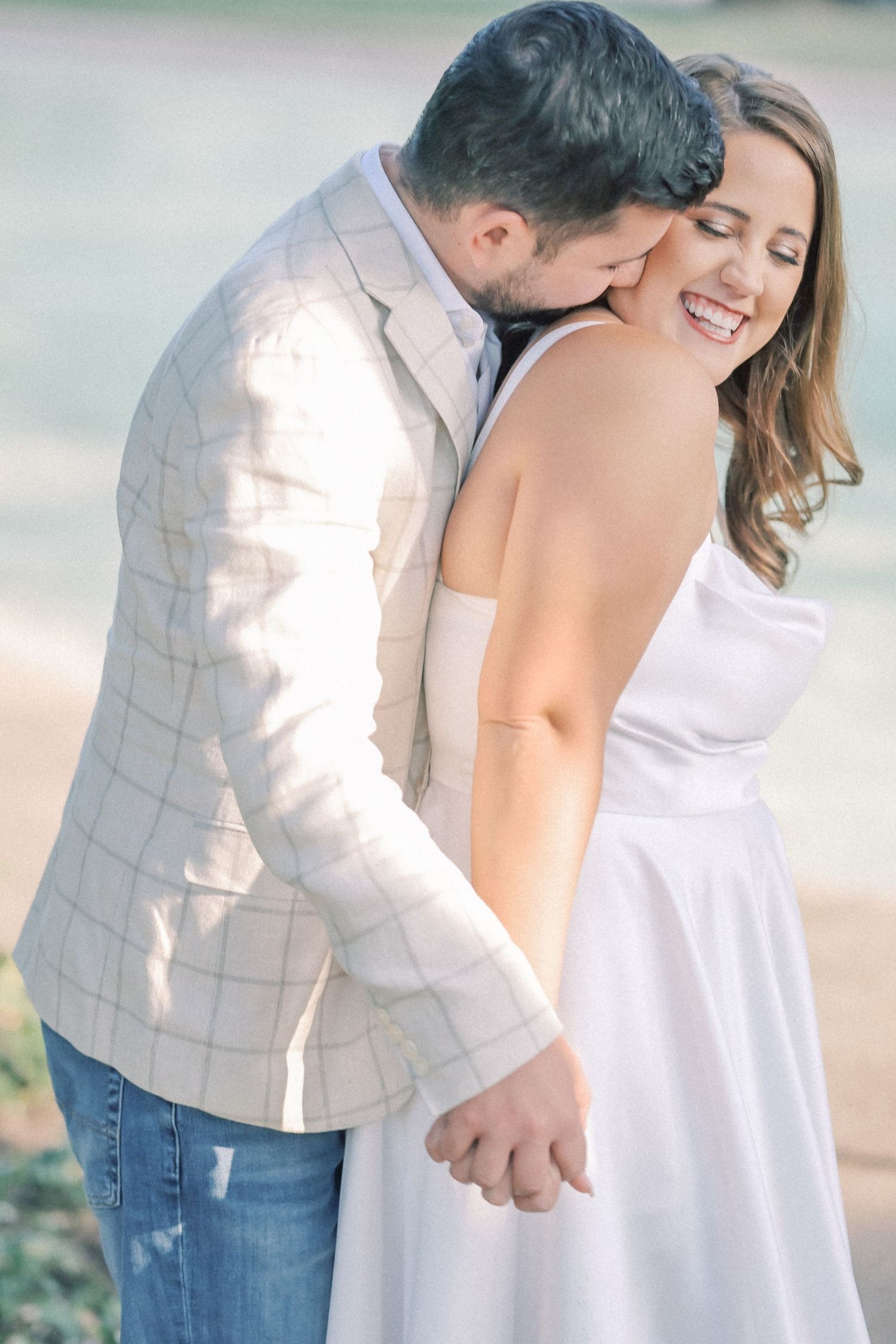 Man in a plaid blazer nuzzles the neck of a laughing woman in a white dress as they hold hands outdoors.