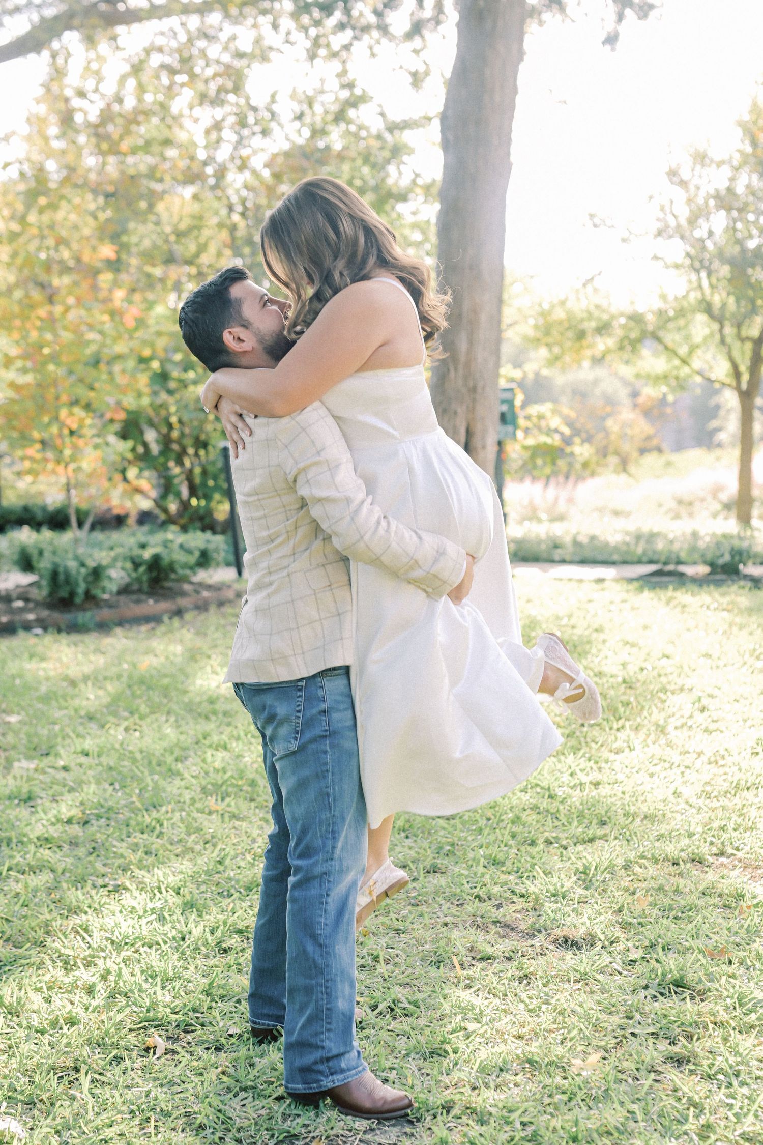 Man in a plaid jacket lifts and kisses a woman in a white dress during an engagement photo session on a sunny, grassy lawn.