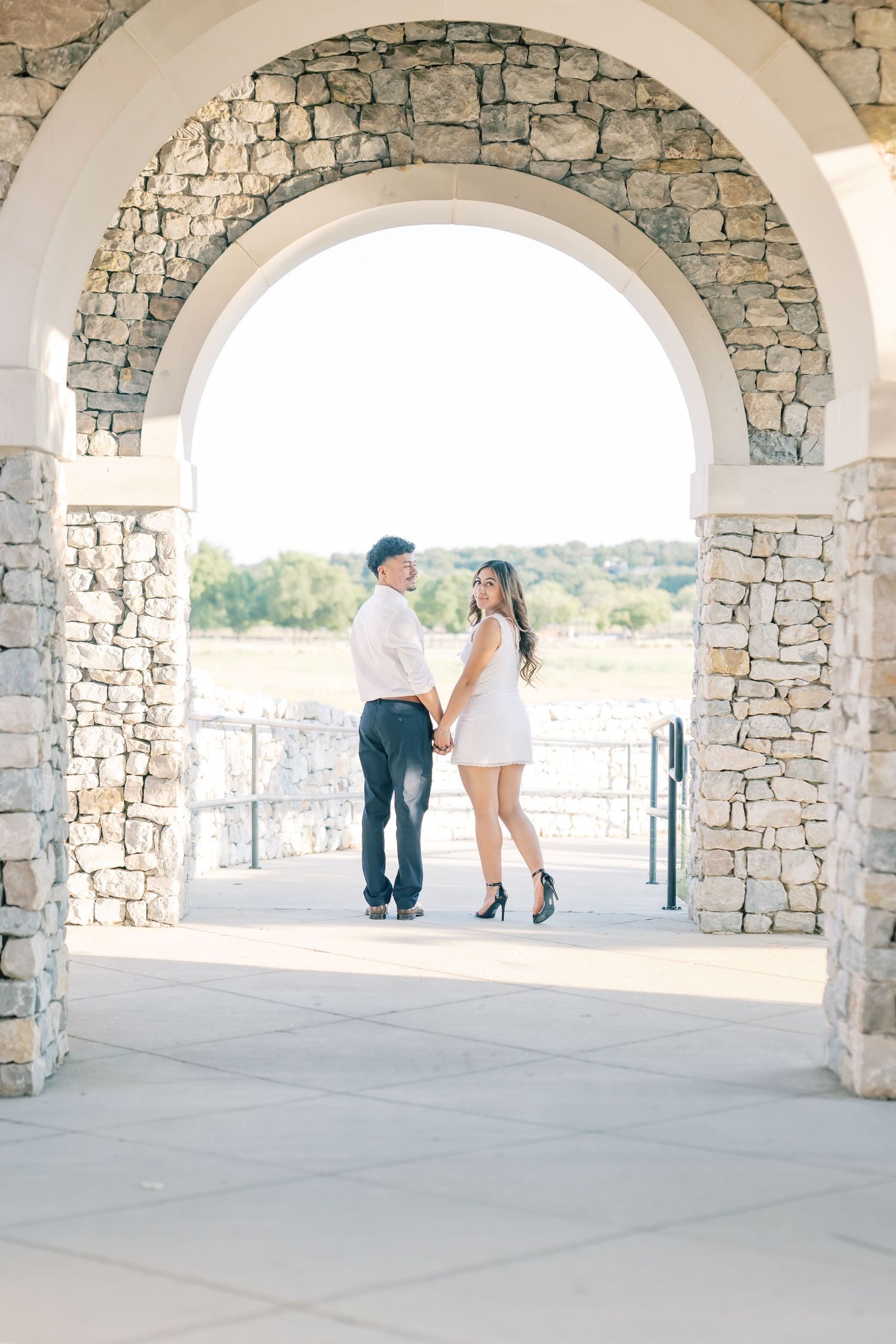 Couple holding hands facing each other under stone archway during golden hour engagement session.