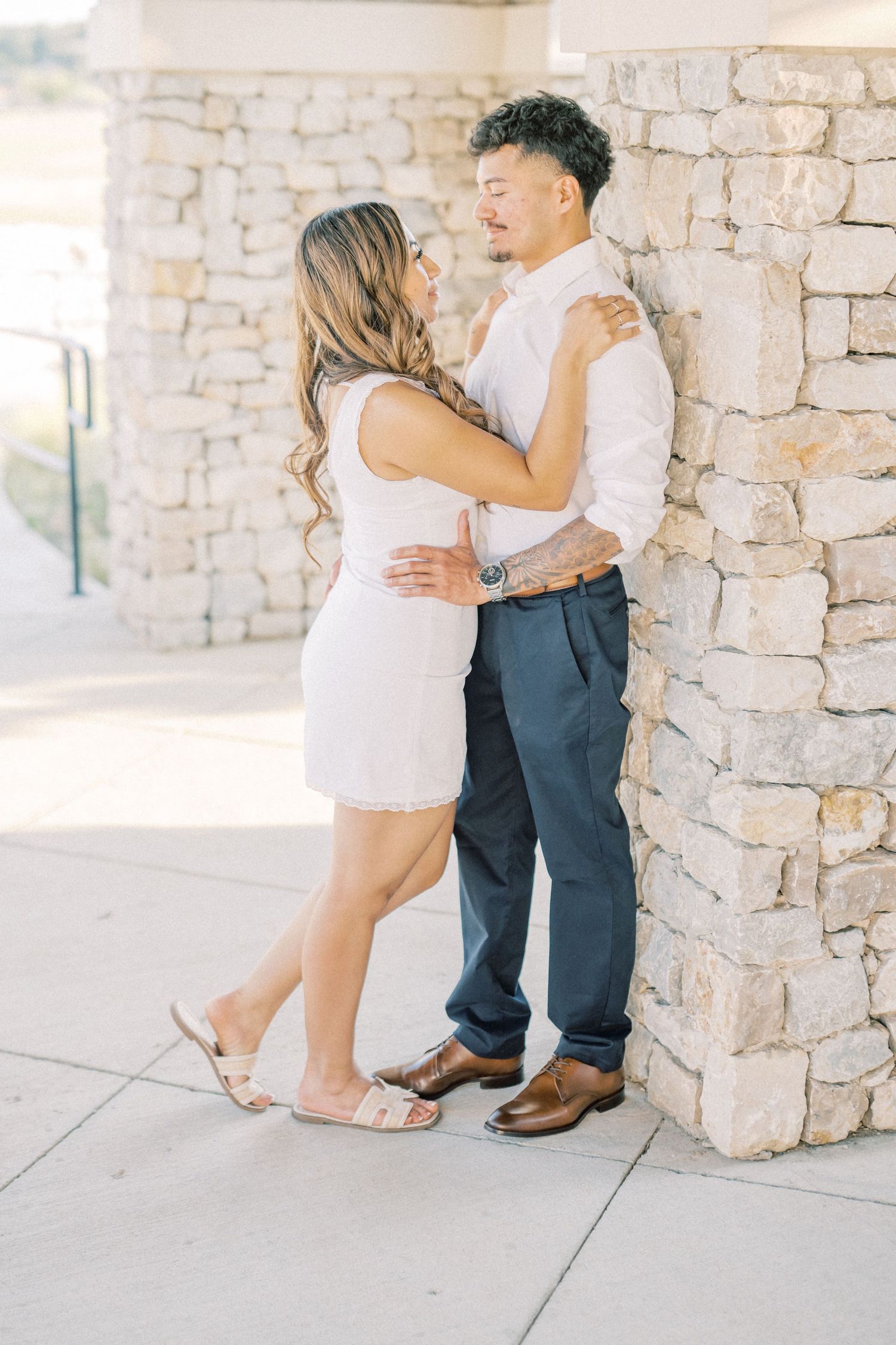Couple embracing against a stone wall, woman in white dress and man in white shirt with navy pants.