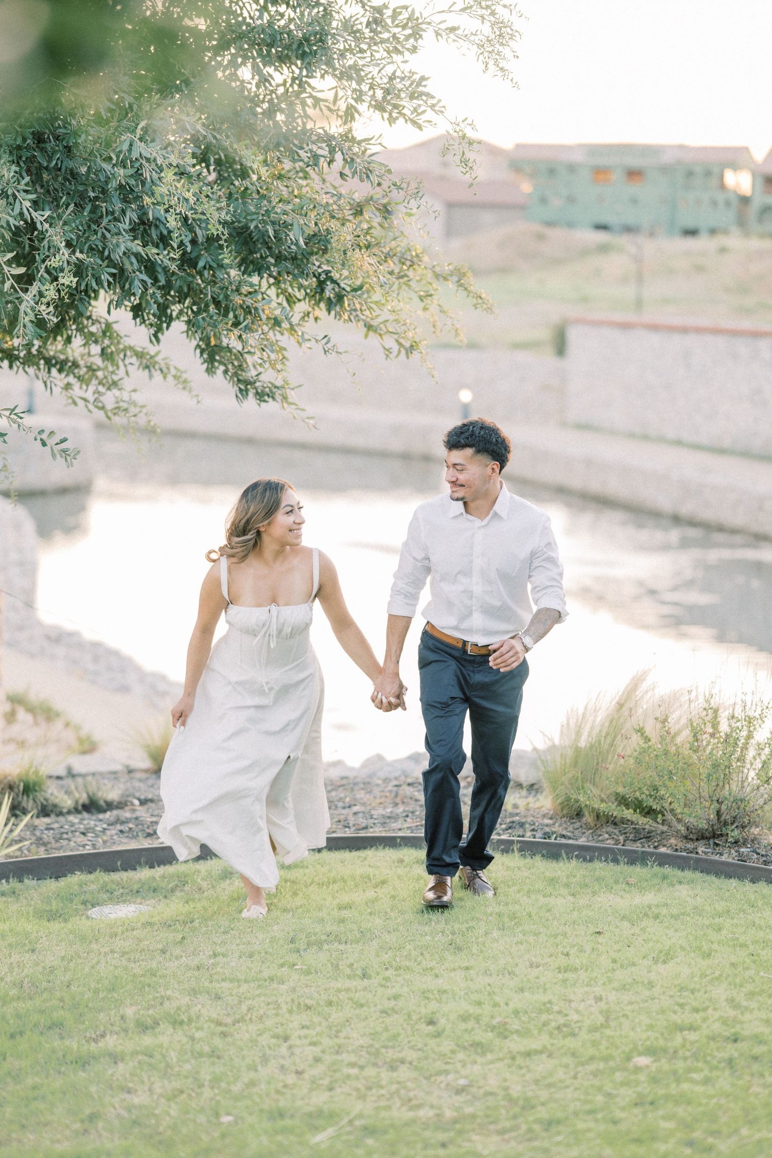 Couple holding hands and walking on grass near a pond during golden hour engagement session.