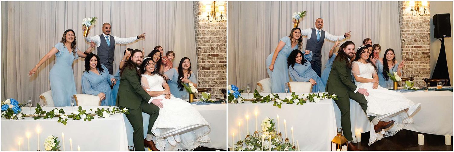 Wedding party poses joyfully at the head table during a elegant reception with floral decorations and soft lighting.