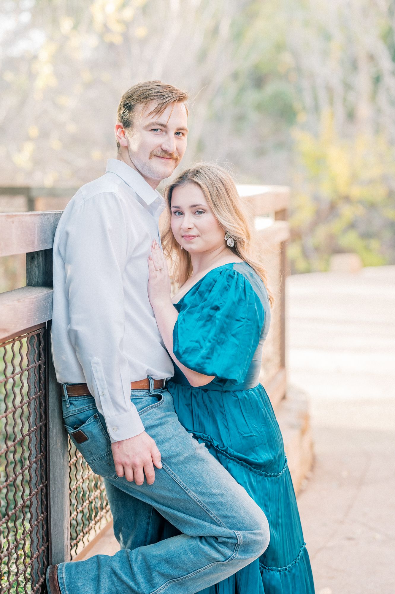 Romantic couple poses together on wooden bridge with woman in teal dress and man in white button down shirt.