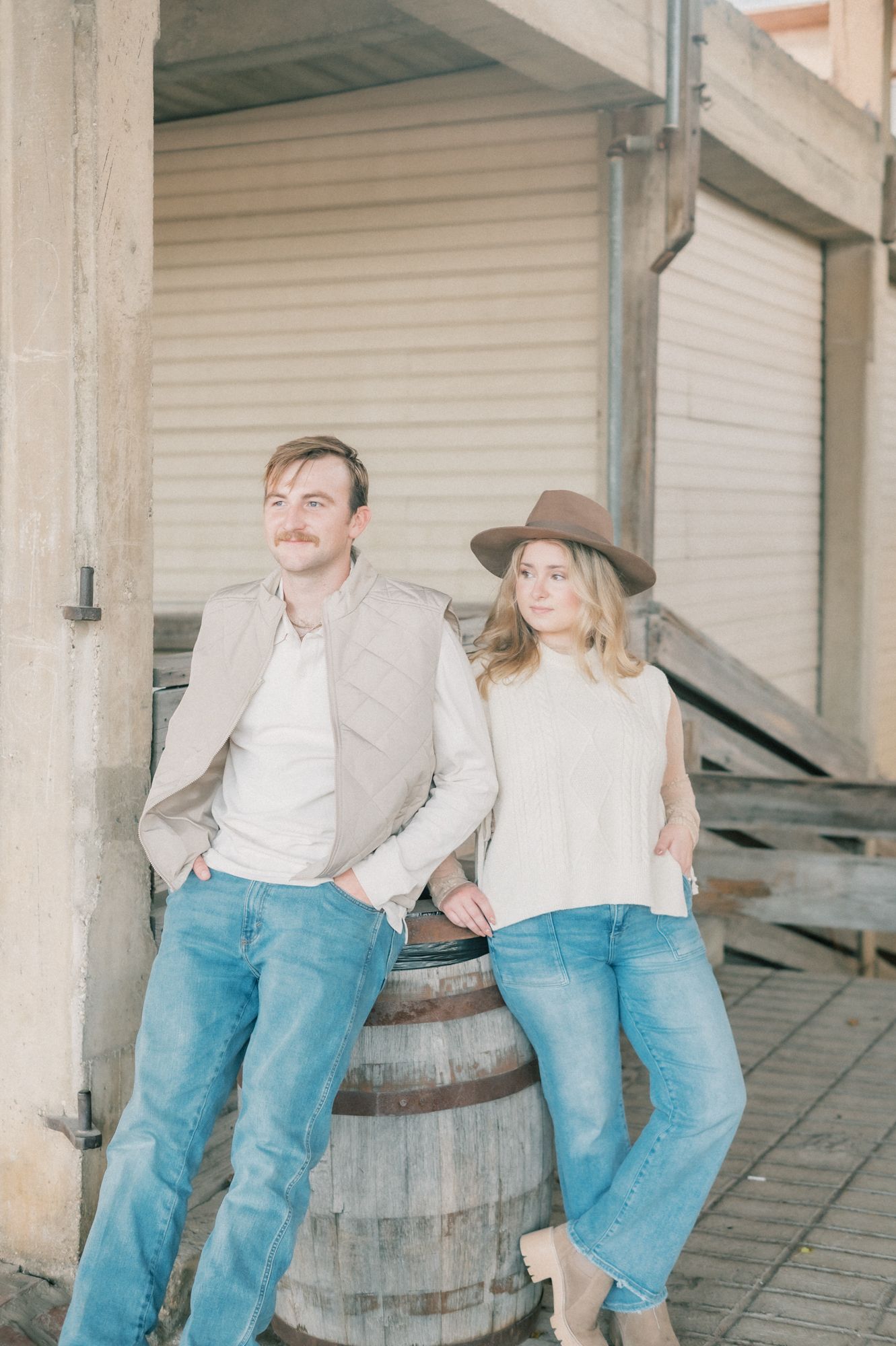 A couple in matching denim and white shirts sits together on a rustic wooden barrel on a porch.
