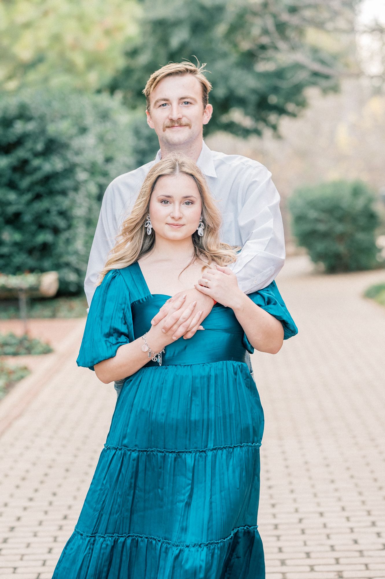 A couple embraces outdoors in a romantic photoshoot, with the person in a teal tiered dress against a natural background.