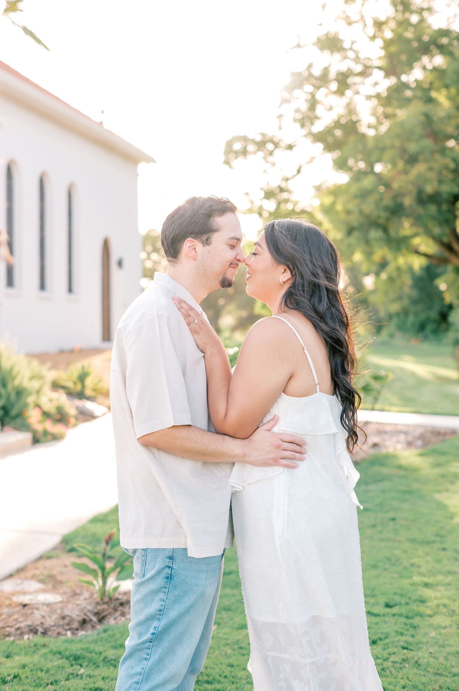A couple embraces outdoors against a sunlit white building with lush greenery in the background.
