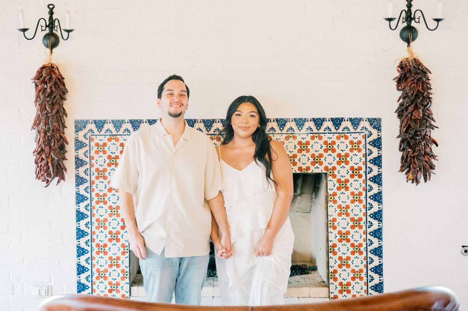 A couple in white and denim poses against a decorative tiled wall with hanging decor and blue bunting.