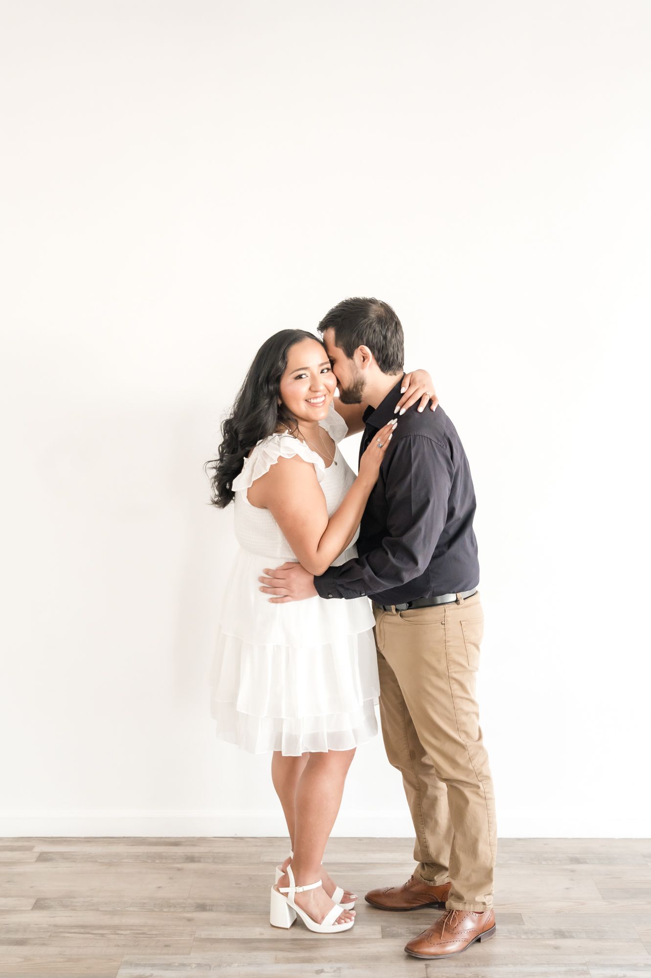 A romantic couple embraces in white and dark attire during an indoor photoshoot.