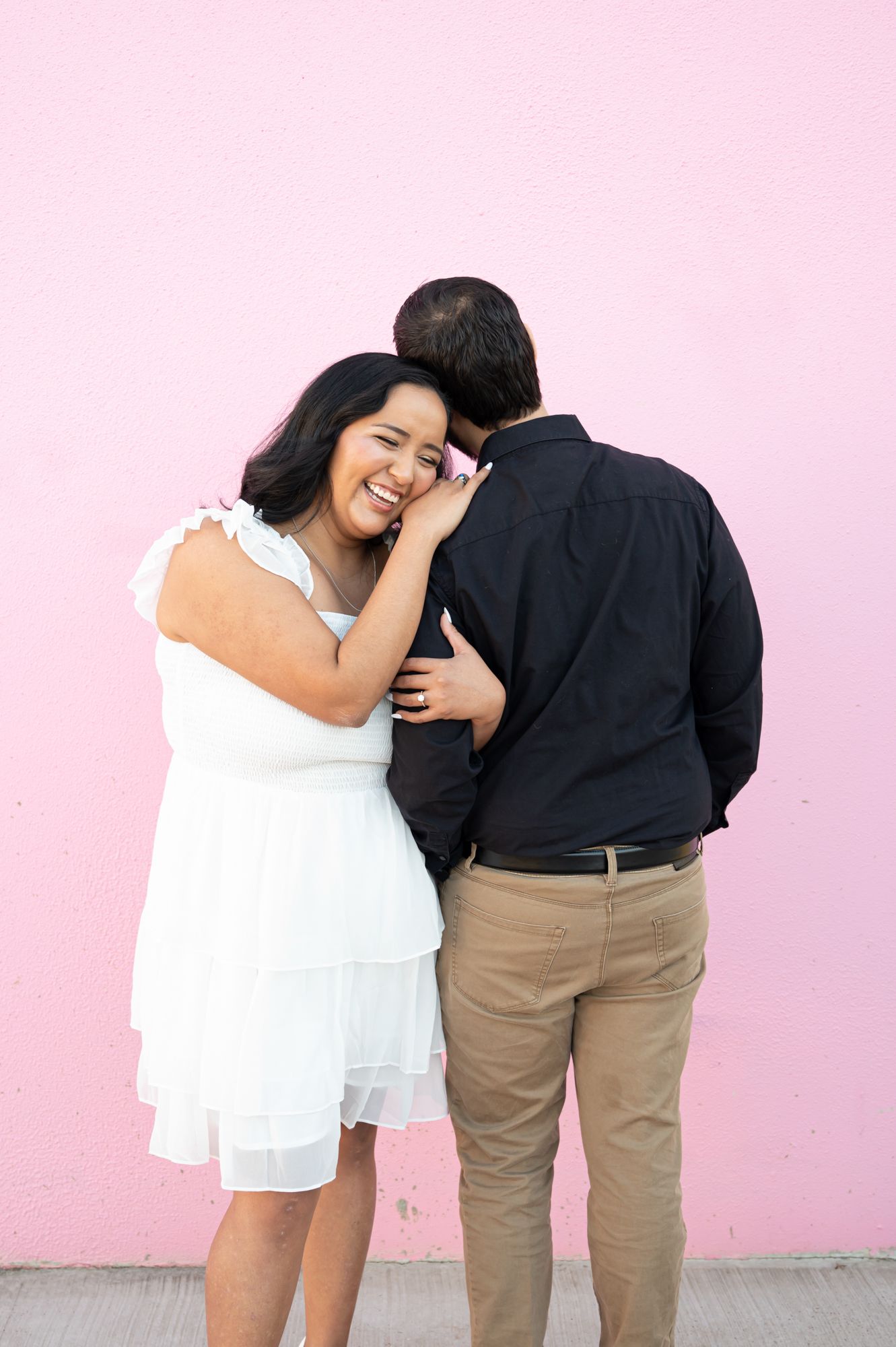 Intimate moment captured between couple in white dress and dark clothing against pink backdrop.