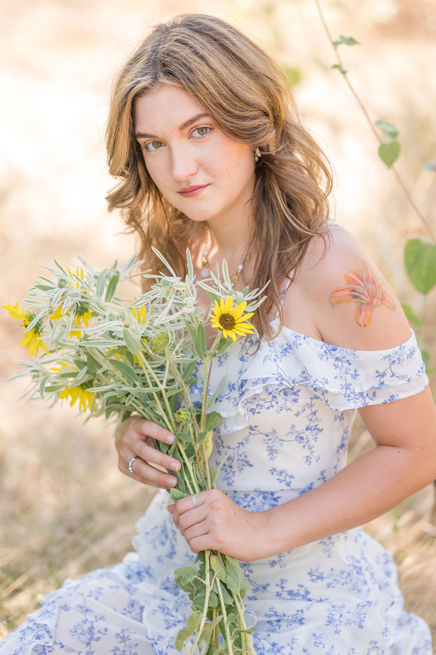 Young woman in floral off-shoulder dress holding wildflowers and sunflowers outdoors, with a butterfly tattoo on her shoulder.