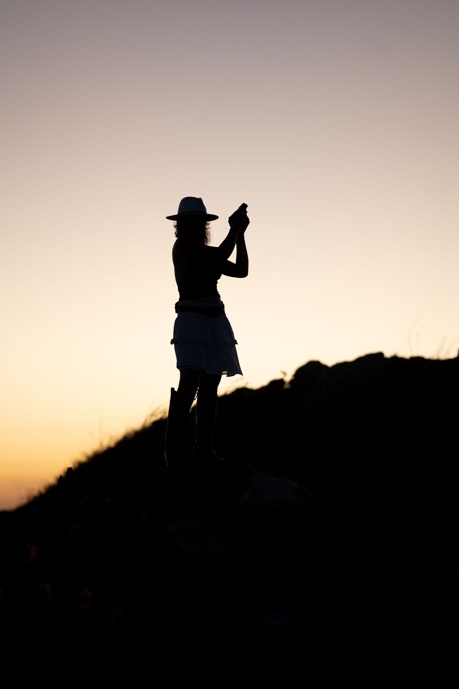 Silhouette of a woman wearing a hat and skirt, standing on a hill taking a photo at sunset.