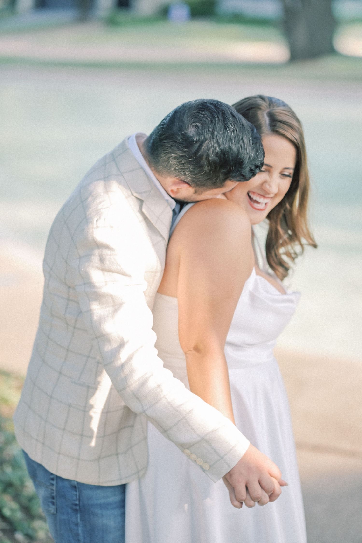 A smiling woman in a white satin dress laughs as a man in a plaid blazer and jeans leans in and kisses her shoulder outdoors.
