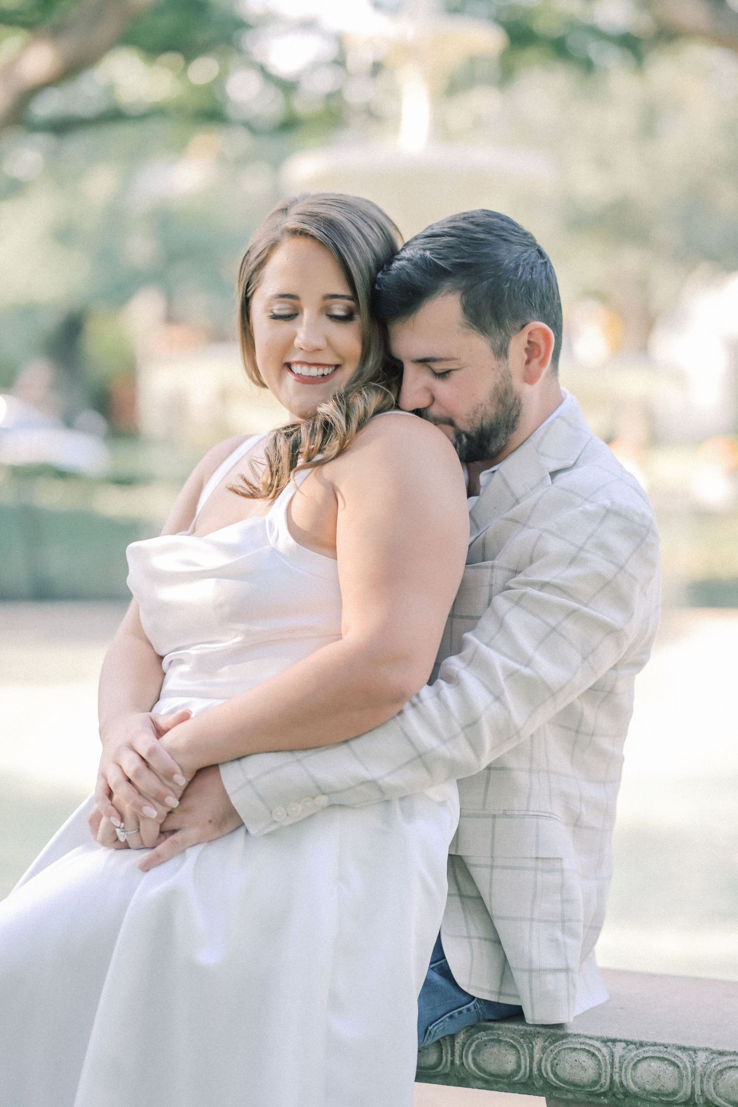 Man in a plaid blazer hugs a smiling woman in a white dress from behind as they sit on a stone bench in a park.