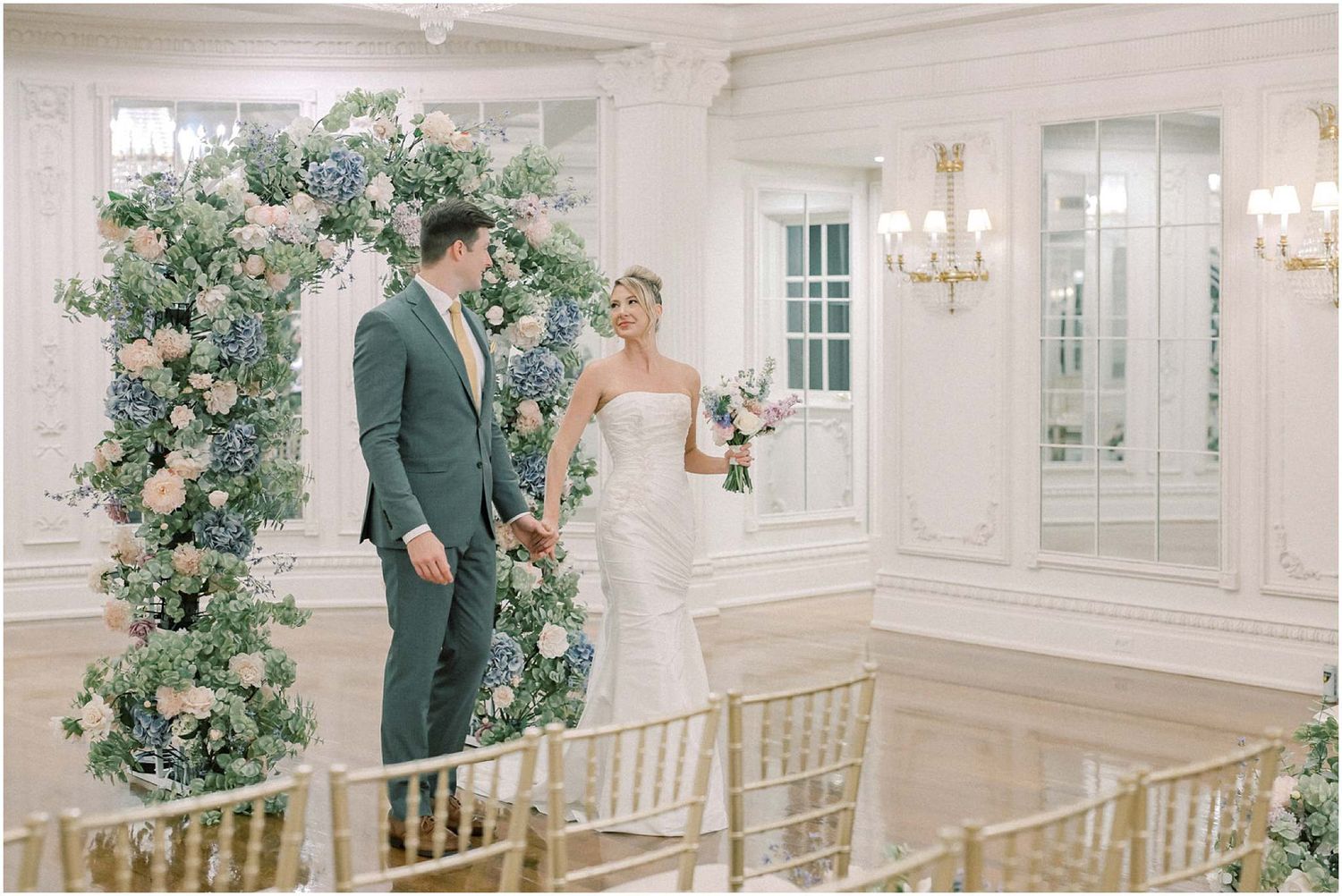 Newlyweds stand together under floral arch in elegant white ballroom with crystal chandeliers.